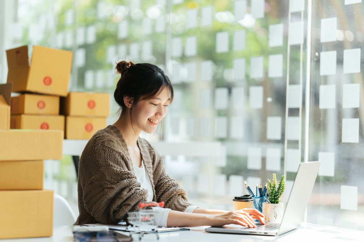 Eine Frau arbeitet am Laptop und im Hintergrund sind mehrere Pakete